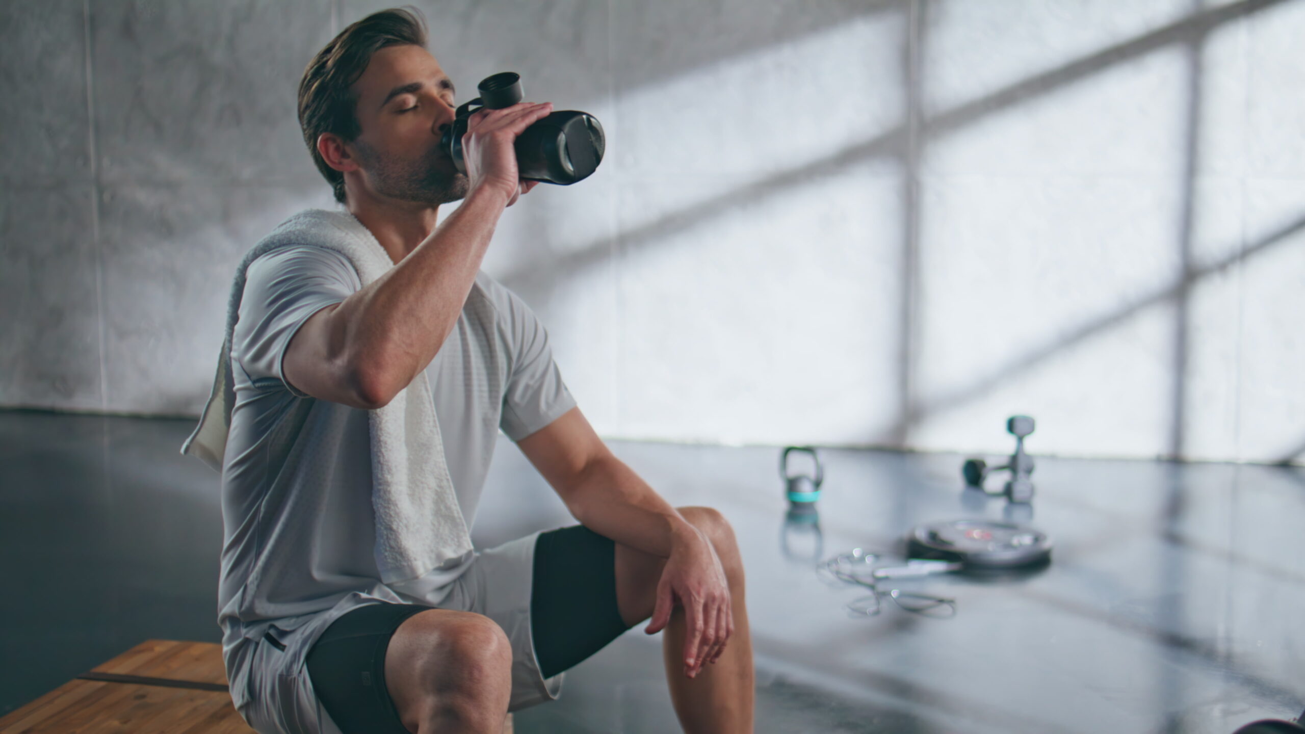 Exhausted man drinking water from sports bottle after fitness training in gym. Male bodybuilder replenishing aqua balance after hard workout. Closeup tired sportsman putting fitness cup on bench